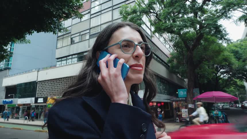 A stylish woman with glasses and a dark coat smiles and laughs while talking on her phone on a busy city street