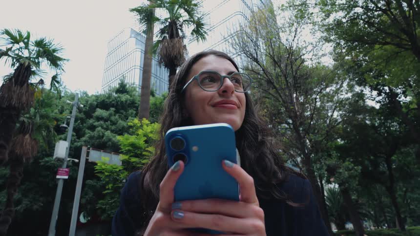 A smiling young businesswoman uses her smartphone while walking through a modern city park, stopping to look up thoughtfully