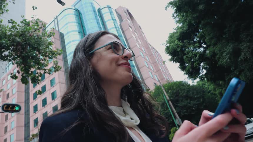 A smiling businesswoman uses her smartphone on a busy city street with modern buildings and trees in the background