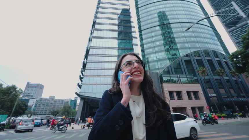 A smiling businesswoman enjoys a phone call on a bustling city street with modern skyscrapers in the background