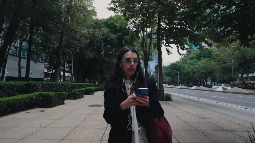 A stylish woman walks in slow motion along a tree-lined city sidewalk, listening to music on her phone as a red bus passes by