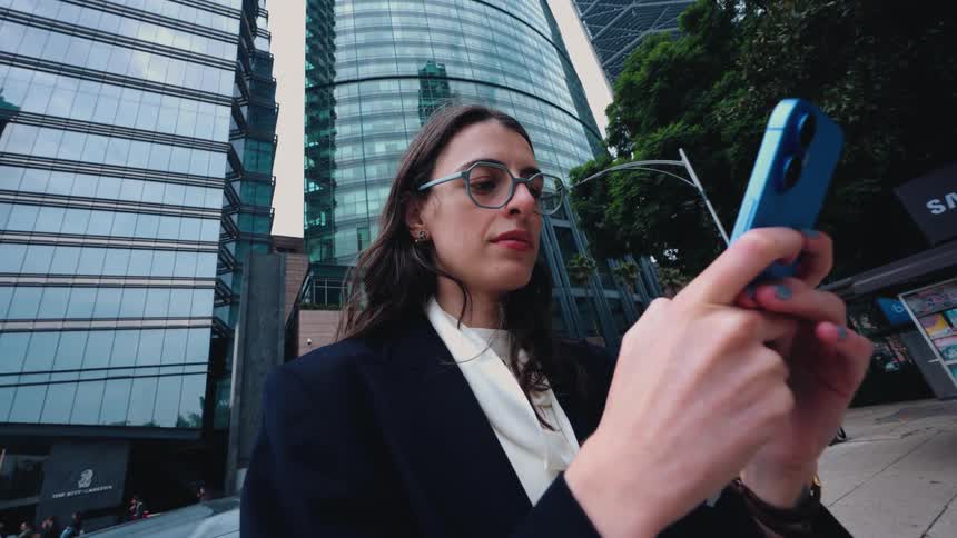 A stylish businesswoman smiles while texting on her smartphone, standing on a bustling city street surrounded by modern glass skyscrapers