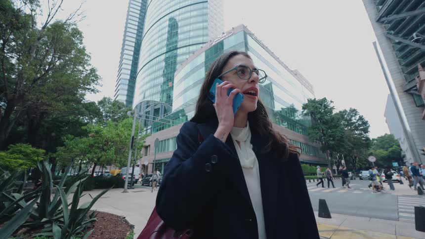 A smiling businesswoman answers her smartphone on a busy city street in front of modern office buildings