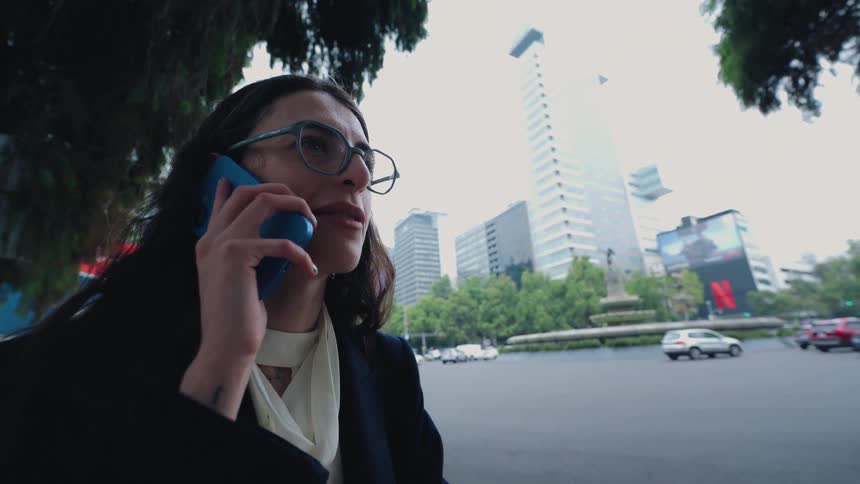 A young businesswoman smiles happily while talking on her smartphone on a busy city street corner with traffic and modern buildings