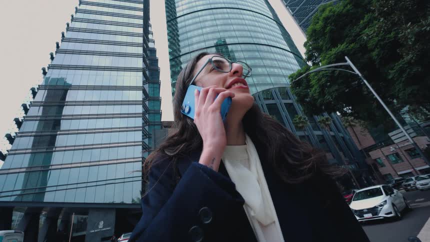 A stylish businesswoman receives good news on a phone call and laughs joyfully on a city street with modern skyscrapers