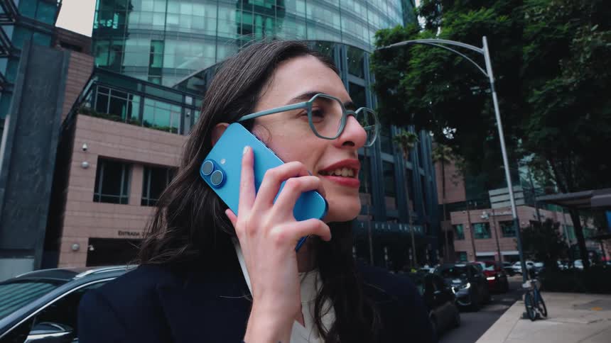 A professional woman in glasses answers her smartphone and walks down a busy city street lined with modern glass buildings