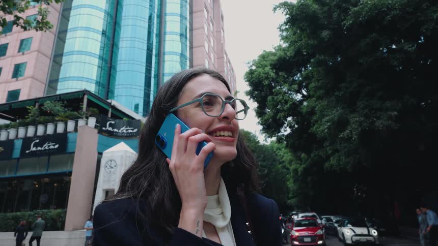 A stylish businesswoman in glasses smiles as she answers her smartphone on a busy city street with modern buildings in the background
