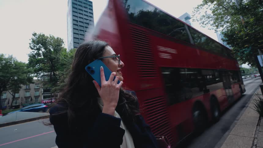 A smiling businesswoman talks on her smartphone on a bustling city avenue as a red double-decker bus passes by in slow motion