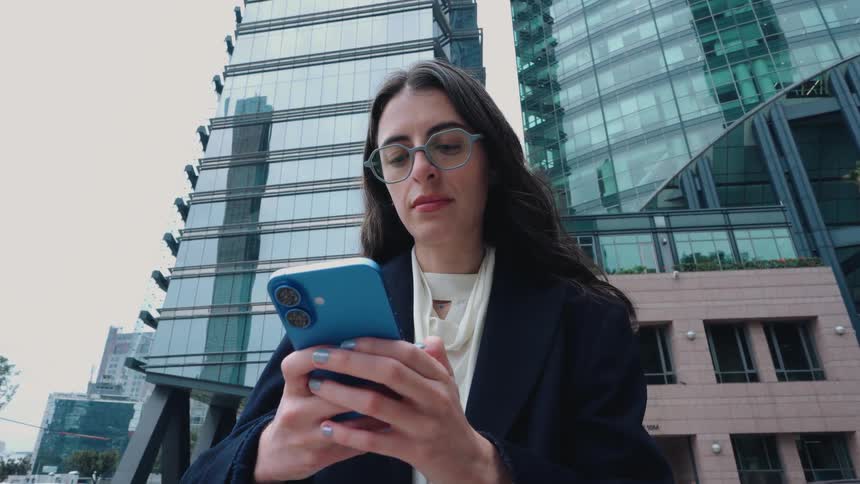A low-angle shot captures a stylish businesswoman in glasses standing in front of a modern glass skyscraper as she focuses on her smartphone