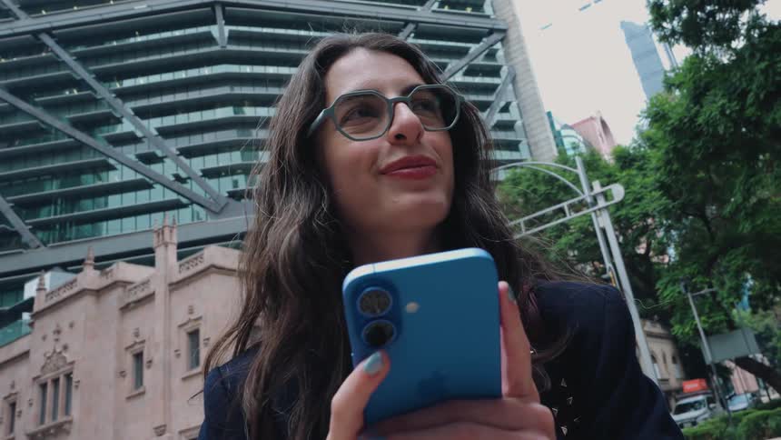A stylish young woman in glasses happily uses her smartphone while standing on a busy city street with modern buildings in the background