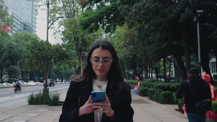 A professional young woman in a blazer smiles while using her smartphone as she walks down a treelined city sidewalk in slow motion