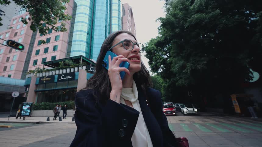 A confident businesswoman with glasses smiles as she answers a phone call on a busy city street corner