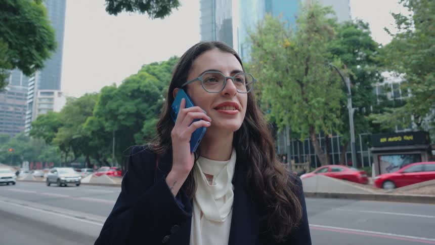 A stylish businesswoman with glasses happily talks on her smartphone while standing on a busy city street with traffic and modern buildings in the background