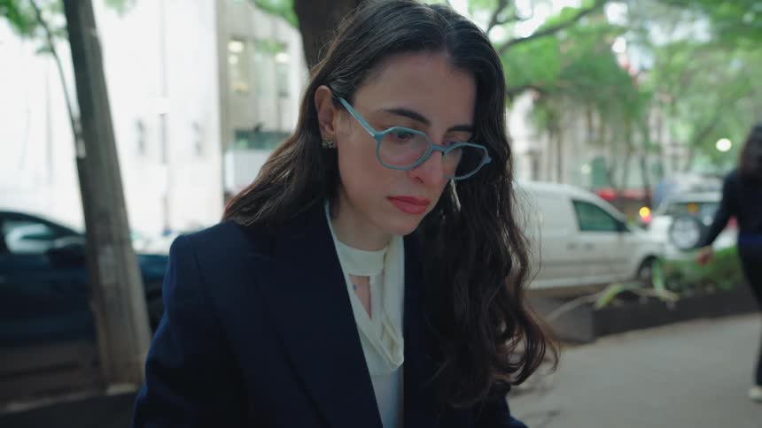 A serious professional woman wearing glasses and a blazer focuses intently on her work while sitting outside on a busy city street