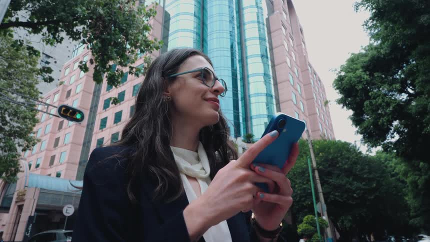 A happy businesswoman in glasses smiles while using her smartphone on a busy city street in front of a modern high-rise building