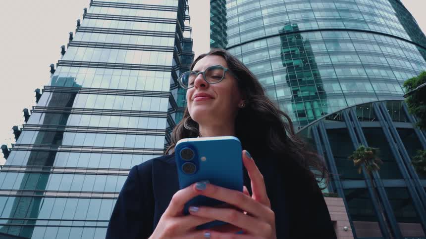 A successful young businesswoman smiles thoughtfully while using her smartphone against a backdrop of towering modern skyscrapers