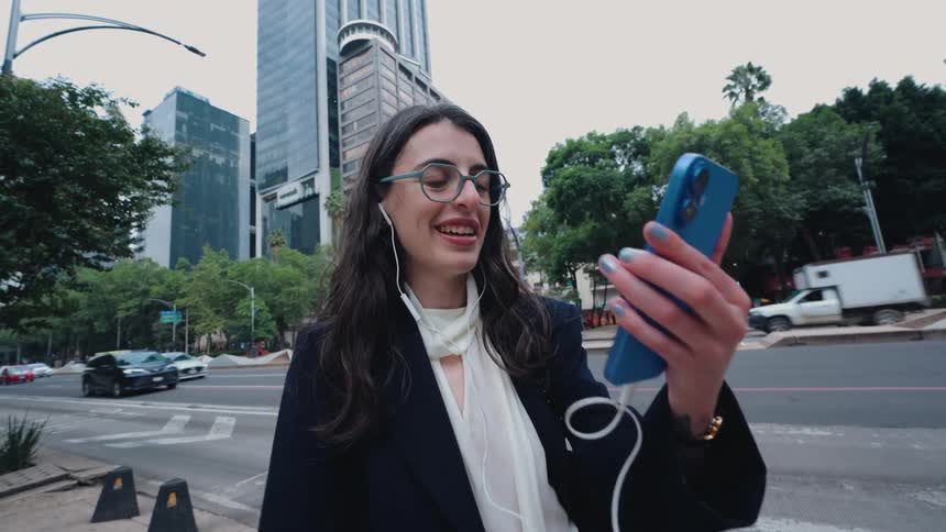 Stylish young businesswoman smiles while on a video call with earphones on a busy city street with skyscrapers in the background