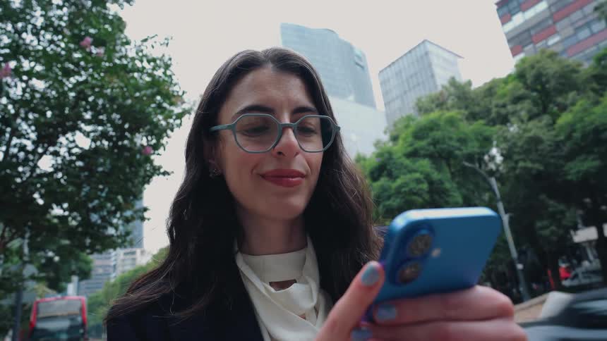 Stylish businesswoman smiles while using her smartphone as she walks along a busy city street with traffic and modern buildings in the background