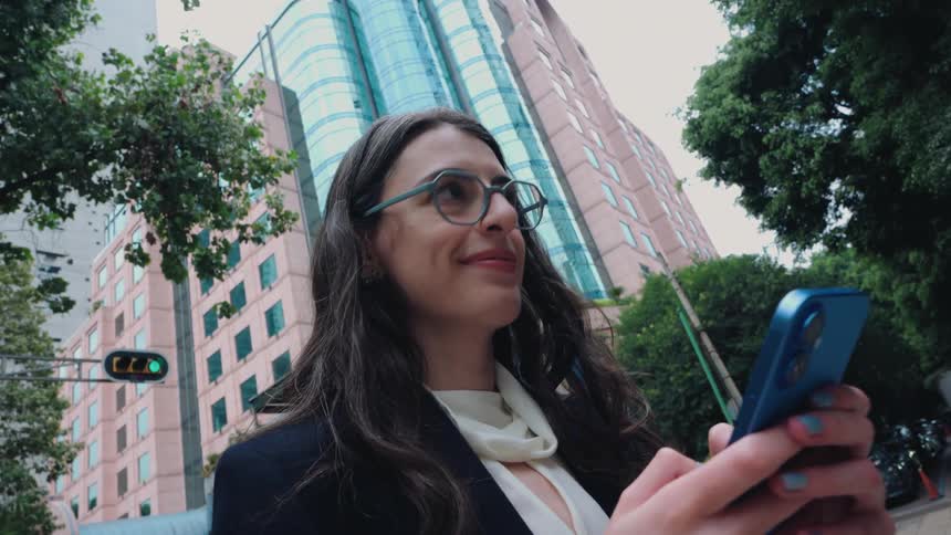 A low angle view shows a happy businesswoman walking in the city and smiling while using her smartphone