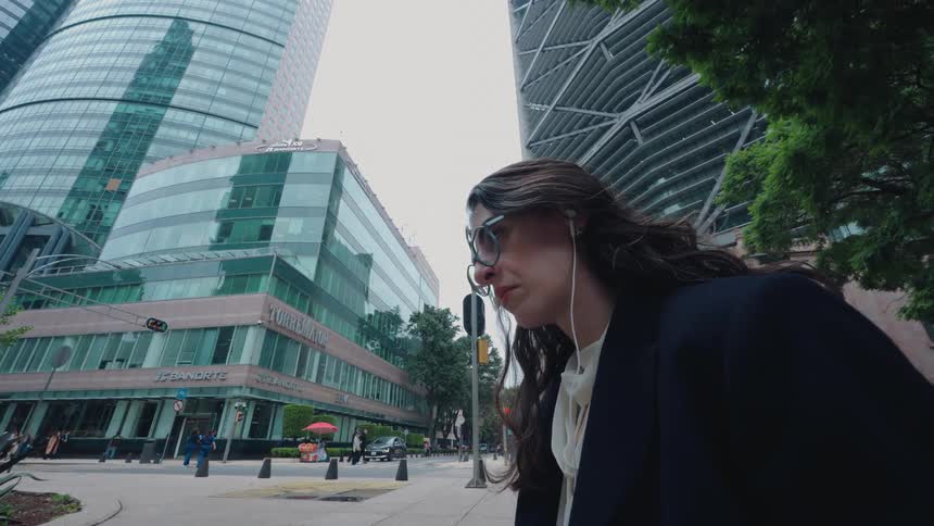 A thoughtful businesswoman with earphones pauses on a city street, looking up at the surrounding modern skyscrapers