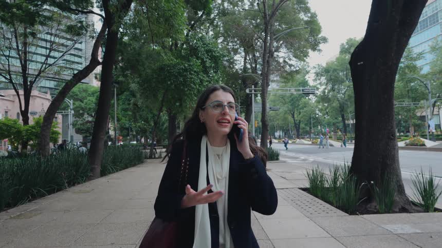 A professional woman in glasses and a coat has an expressive conversation on her smartphone while walking down a tree-lined city sidewalk in slow motion