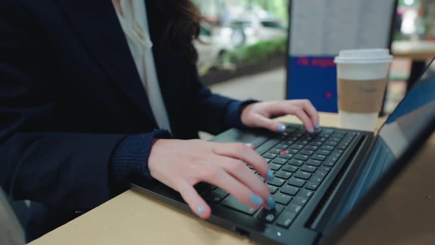 Close-up shot of a woman with blue nail polish typing on a laptop keyboard while working remotely at a cafe
