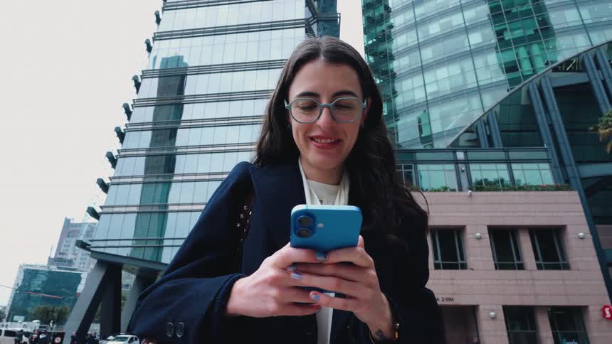A smiling businesswoman happily texts on her smartphone while standing in front of a modern glass skyscraper in the city