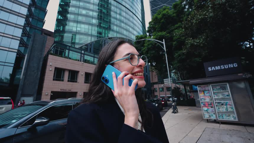 A young businesswoman with glasses talks on her smartphone while standing on a busy city street in front of a modern skyscraper