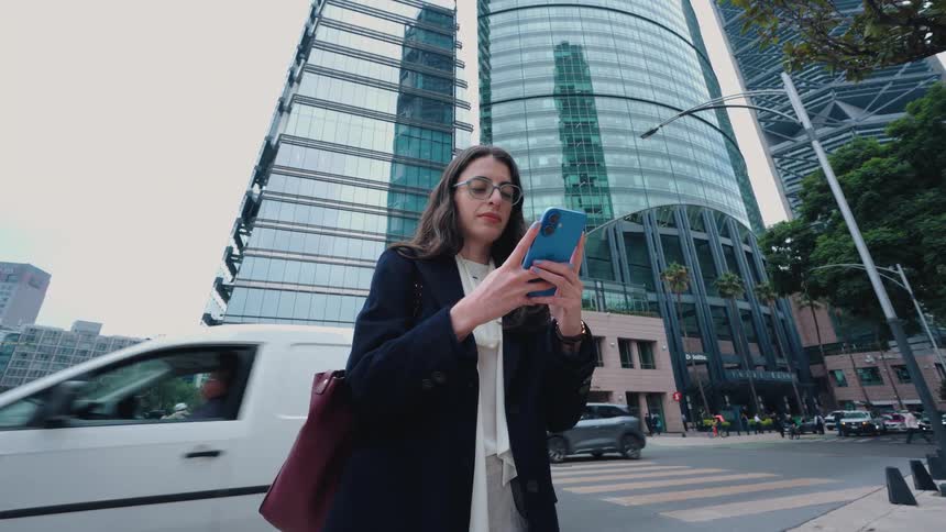 A professional woman in a blazer and glasses uses her smartphone on a bustling city corner with a modern glass skyscraper behind her