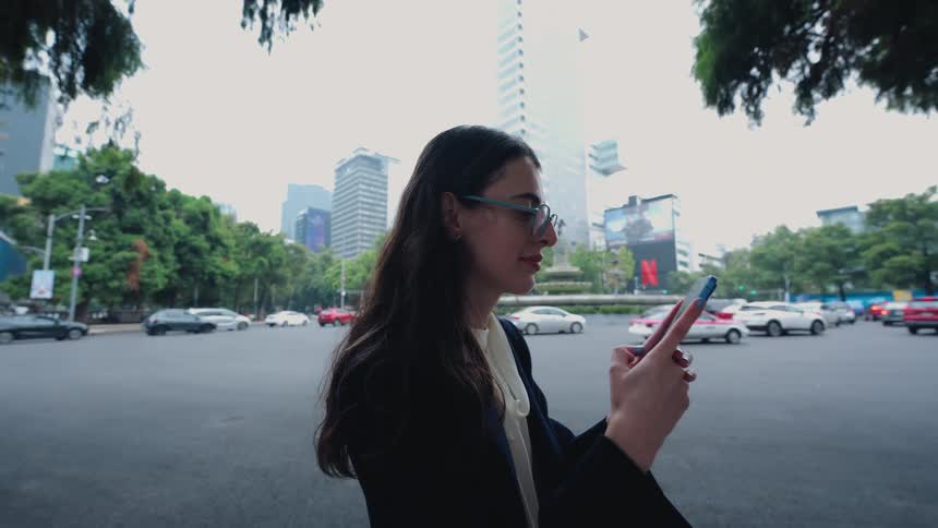 A smiling businesswoman uses her smartphone on a busy street corner with city traffic passing in the background