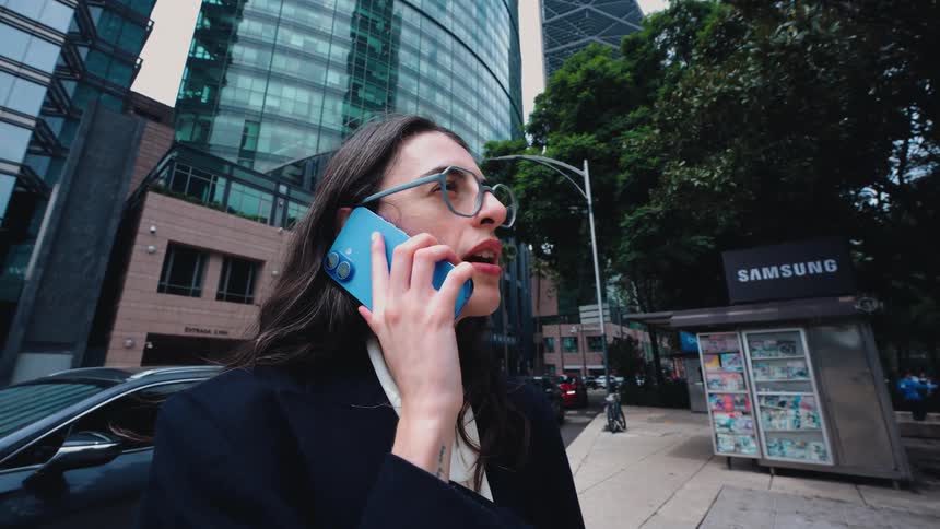 A stylish businesswoman smiles happily while taking a phone call on a busy street in a modern city