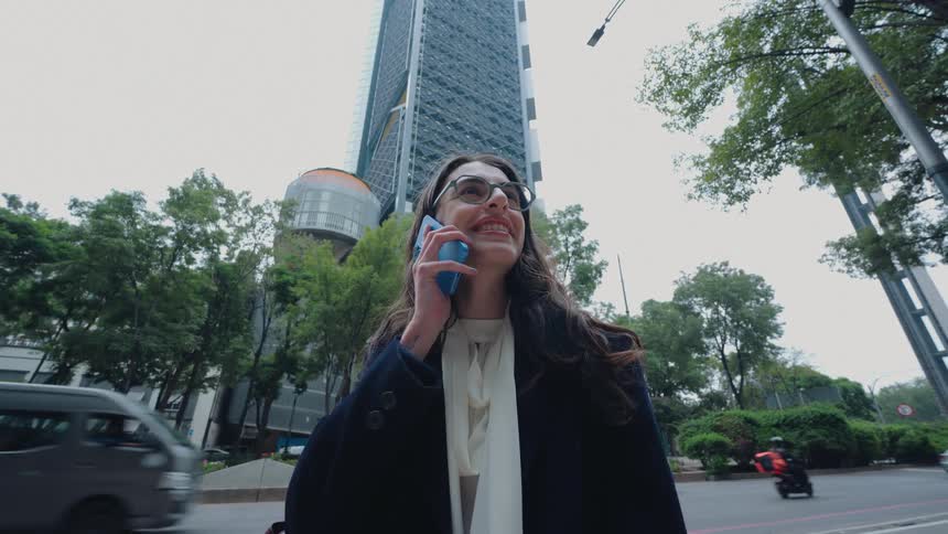 Stylish young woman happily answers a phone call while standing on a bustling city street next to a modern skyscraper