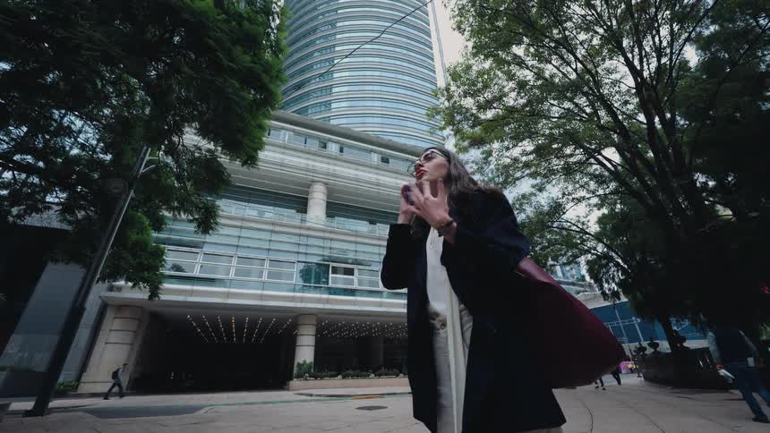A low-angle, slow-motion view shows a businesswoman having a serious phone conversation, then smiling as she walks past a modern skyscraper