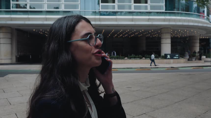 A businesswoman in glasses talks on her smartphone in slow motion while standing on a city sidewalk in front of a modern building