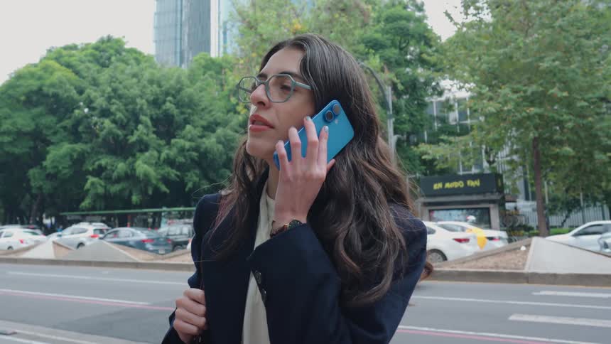 An elegant young woman makes a phone call on a bustling city street, smiling as she talks amidst traffic and skyscrapers