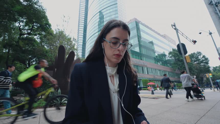 A young businesswoman in glasses and earphones sits on a busy city sidewalk, focused on her smartphone as people pass by in slow motion