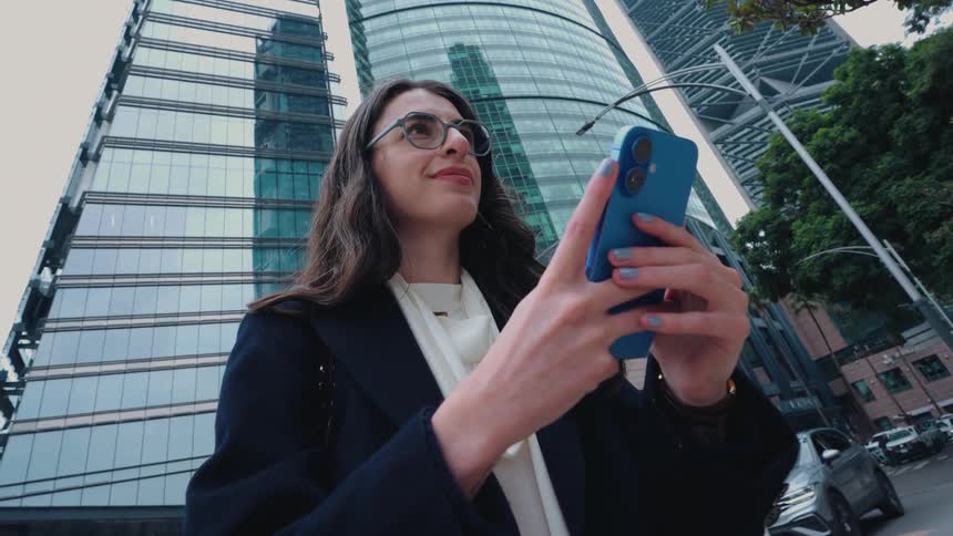 A stylish businesswoman uses her smartphone on a bustling city corner with modern glass skyscrapers in the background
