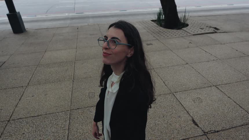 High-angle slow-motion shot of a businesswoman with glasses stopping on a city sidewalk and looking up with a smile as cars pass by
