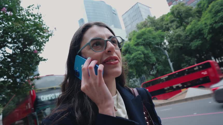 A businesswoman wearing glasses and a blazer talks on her phone while standing on a busy city street with skyscrapers in the background
