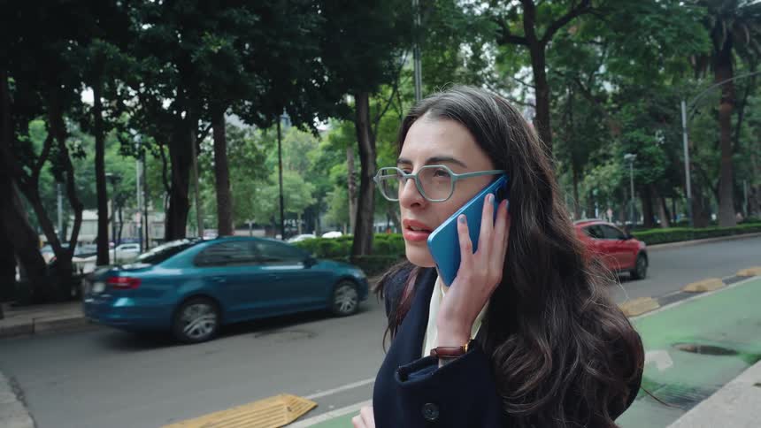 A stylish businesswoman in glasses talks on her phone on a city street, her expression changing from serious to a happy smile in slow motion