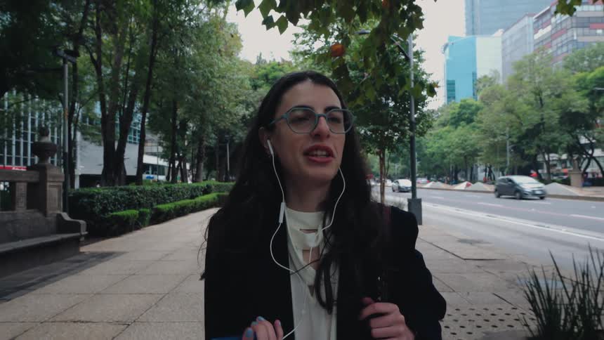 A businesswoman wearing glasses and earbuds talks on a video call while walking along a busy city street in slow motion