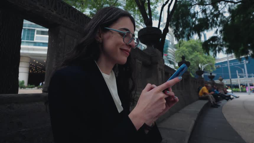 A smiling young businesswoman in glasses sits on a stone ledge in a city park, happily scrolling through her smartphone
