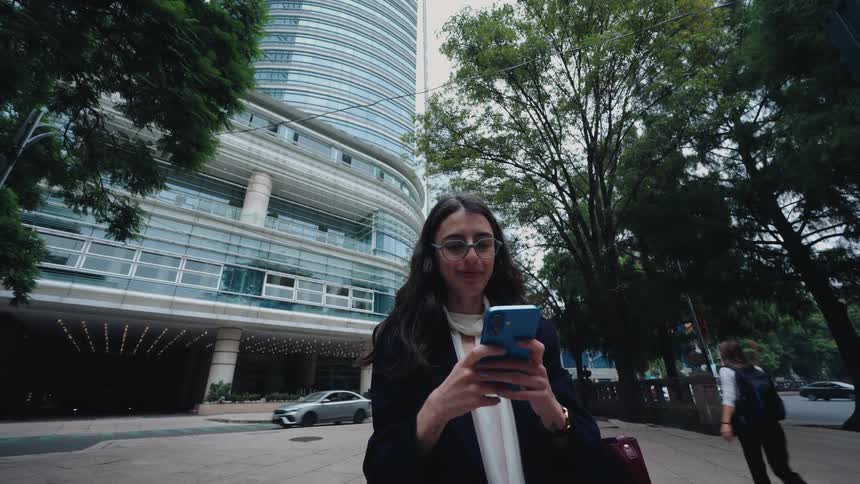 A stylish businesswoman in glasses uses her smartphone while waiting on a city sidewalk in front of a modern skyscraper
