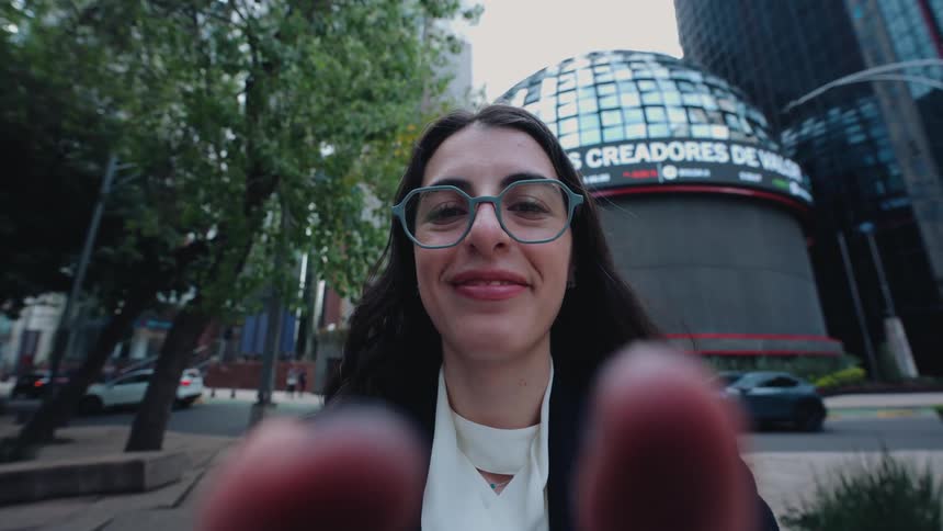 A smiling woman in professional attire holds the camera for a POV selfie video on a city street in front of a modern financial building