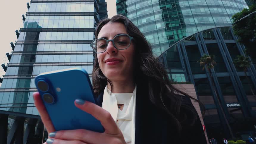 A low-angle view shows a smiling businesswoman using her smartphone while standing in a modern city financial district