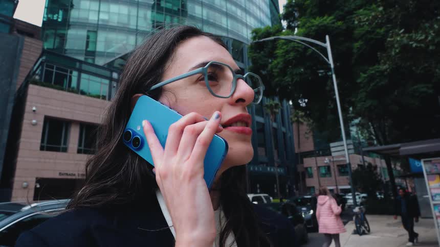 A professional businesswoman in glasses takes an important phone call while standing on a bustling city street with modern skyscrapers in the background