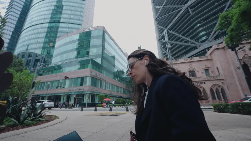 A young businesswoman listens to music with earphones while working on her laptop on a city street surrounded by modern skyscrapers