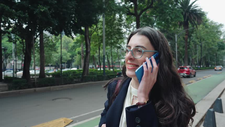 A professional woman in glasses smiles while having a pleasant phone conversation on a bustling city street lined with trees