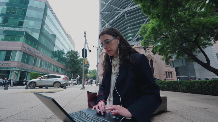 A professional woman works on her laptop and smartphone on a bench in a bustling financial district
