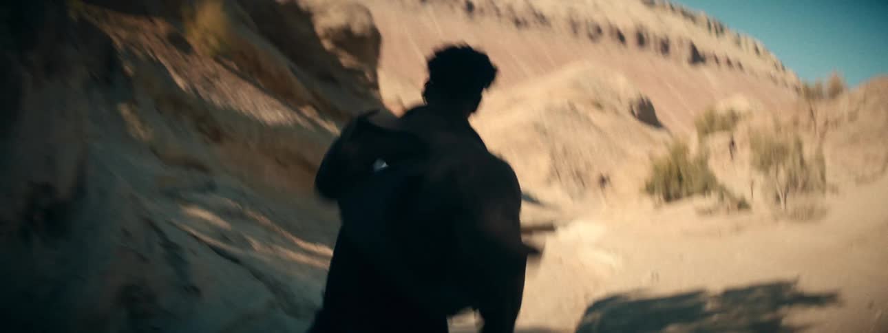 An urgent, shaky handheld shot follows a man as he runs through a dry desert canyon during the day